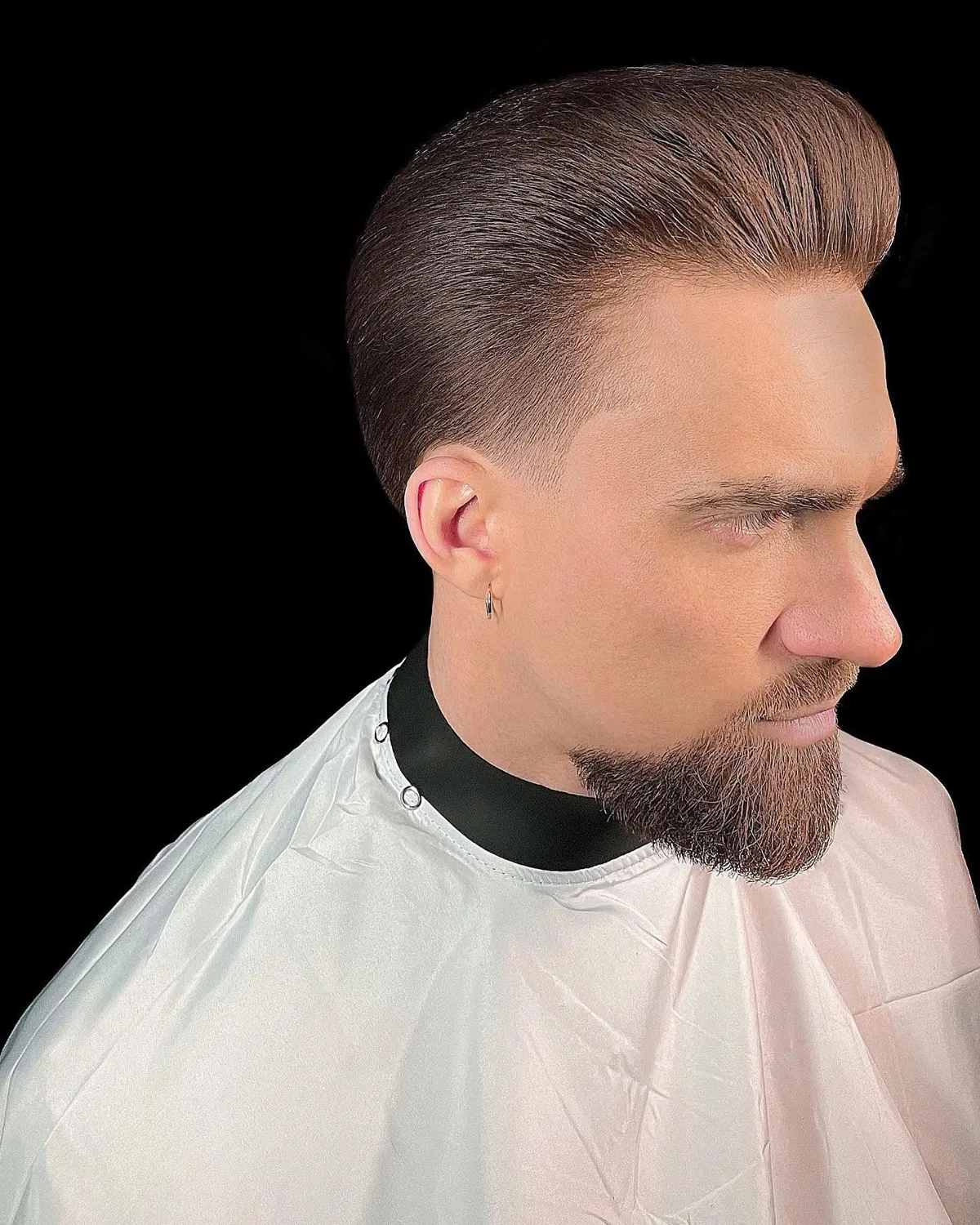 Vīrietis profilā ar pompadour griezumu, taper fade pāreju un koptu bārdu uz melna fona
